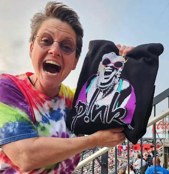 Actress from Marvelous Mrs. Maisel wearing glasses and tie-dye shirt, holding a colorful P!nk sweatshirt outdoors. Actress from Marvelous Mrs. Maisel wearing glasses and tie-dye shirt, holding a colorful P!nk sweatshirt outdoors.