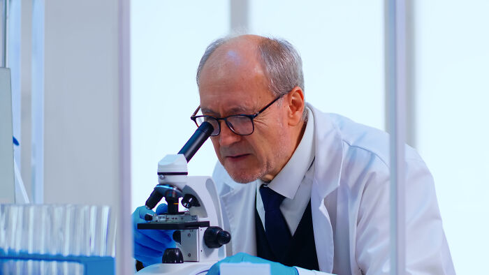 Older male scientist wearing glasses and gloves looking through microscope in a lab, highlighting disturbing science facts.