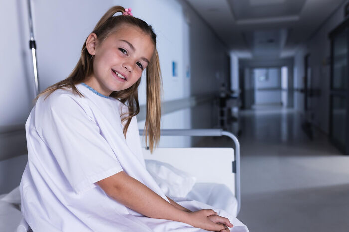 Young girl in hospital gown sitting on a bed, smiling in a quiet hallway, men missed delivery babies concept.