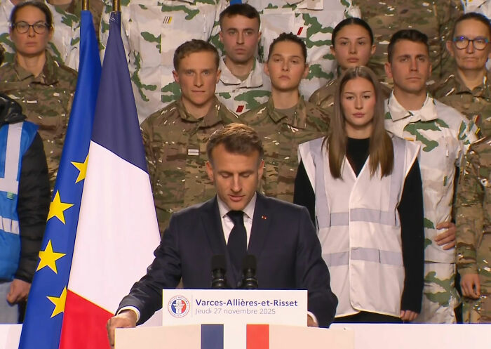 French soldier standing behind Macron during speech, displaying odd behavior as France faces rising tensions and conspiracy theories.