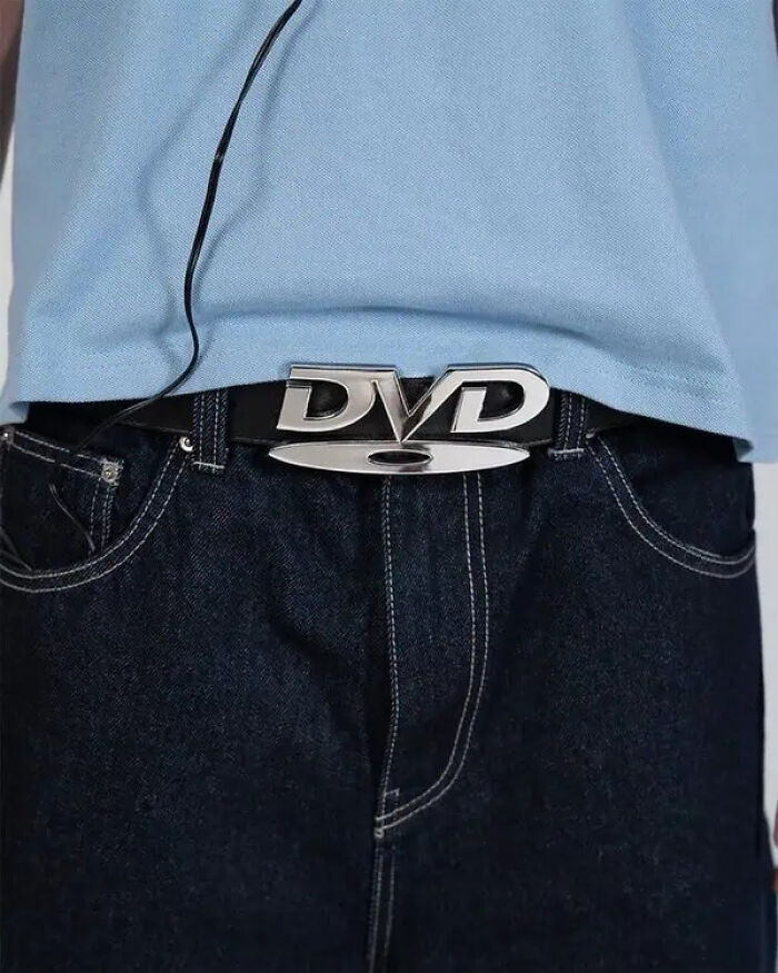 Close-up of a person wearing dark jeans and a belt with a trippy DVD logo buckle, evoking a surreal fashion style.