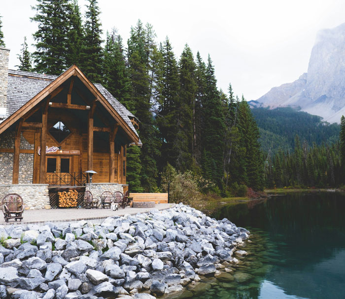 Rustic lakeside cabin surrounded by tall pine trees and mountains, reflecting calm water and rocky shore. Rustic lakeside cabin surrounded by tall pine trees and mountains, reflecting calm water and rocky shore.
