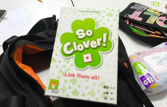 So Clover party game box on a black backpack with colorful school supplies nearby on a white table.