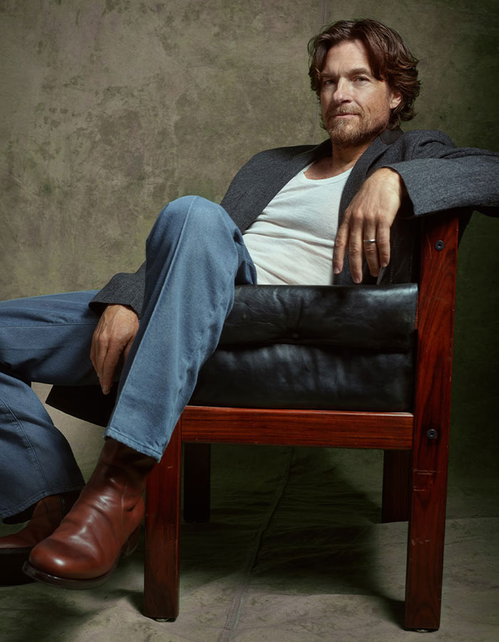 Jason Bateman sitting relaxed in a chair, wearing a gray blazer and brown boots, raising eyebrows with a remark about sister Justine Jason Bateman sitting relaxed in a chair, wearing a gray blazer and brown boots, raising eyebrows with a remark about sister Justine