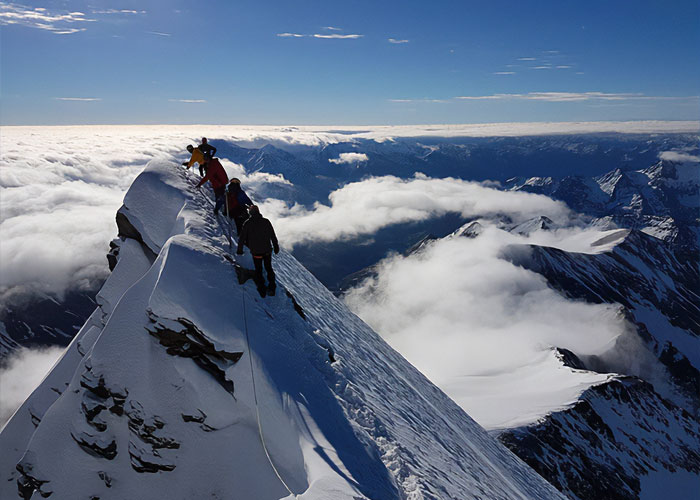 Climbers ascend a steep snowy mountain ridge in Austria, linked by ropes against a backdrop of clouds and peaks.