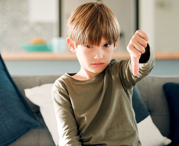 Young boy with serious expression sitting on couch, giving a thumbs down gesture, showing next-level evil attitude.