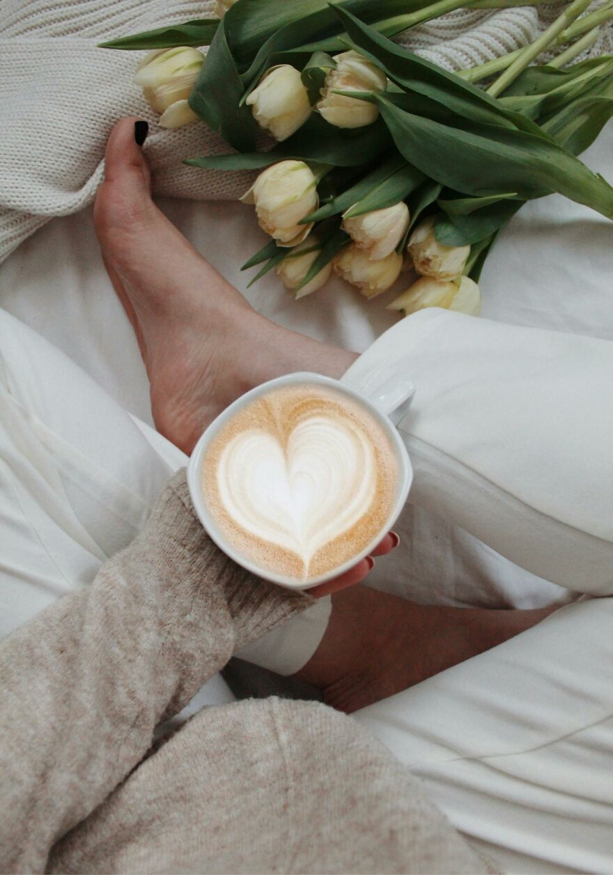Person holding a cup of coffee with heart-shaped foam art near yellow tulips, cozy setting for good morning message to my love. Person holding a cup of coffee with heart-shaped foam art near yellow tulips, cozy setting for good morning message to my love.