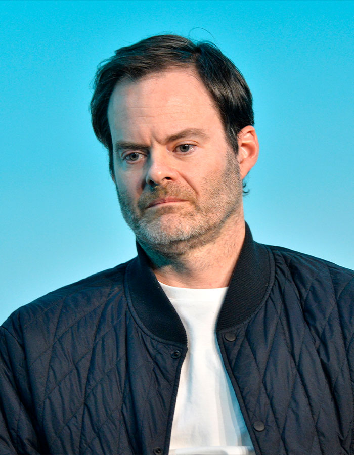 Man with a serious expression wearing a black quilted jacket and white shirt against a blue background at a celebrity event Man with a serious expression wearing a black quilted jacket and white shirt against a blue background at a celebrity event