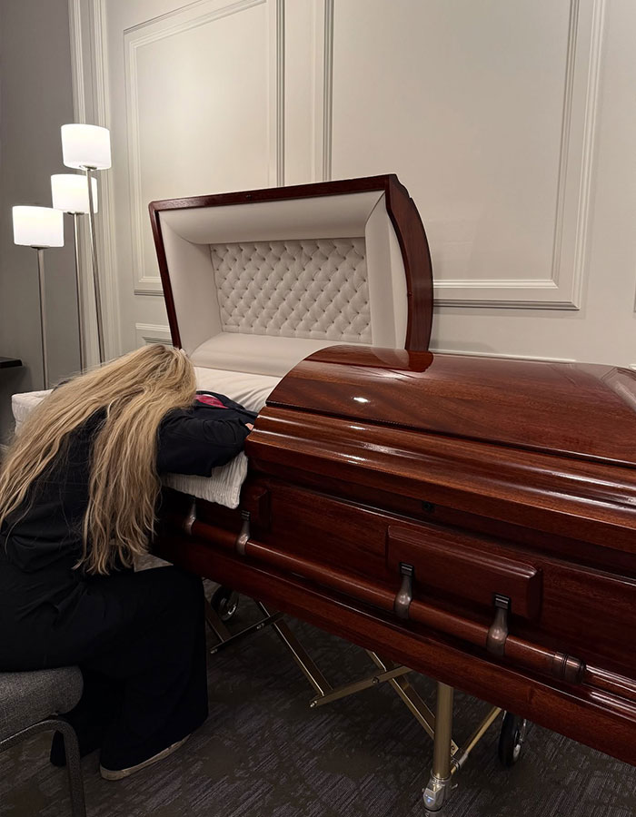 Woman with long blonde hair leans on a polished wooden casket in a subdued room, reflecting on Erika Kirk's curated moment. Woman with long blonde hair leans on a polished wooden casket in a subdued room, reflecting on Erika Kirk's curated moment.