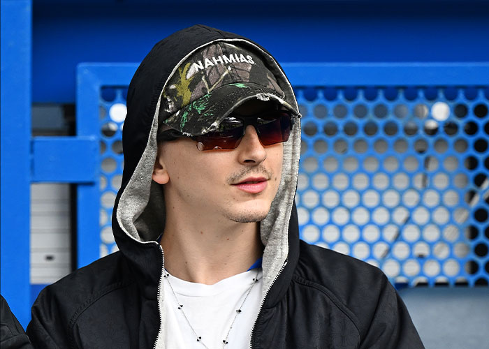 Timothée Chalamet wearing sunglasses and a cap, facing sideways with a casual black hoodie in an outdoor setting. Timothée Chalamet wearing sunglasses and a cap, facing sideways with a casual black hoodie in an outdoor setting.