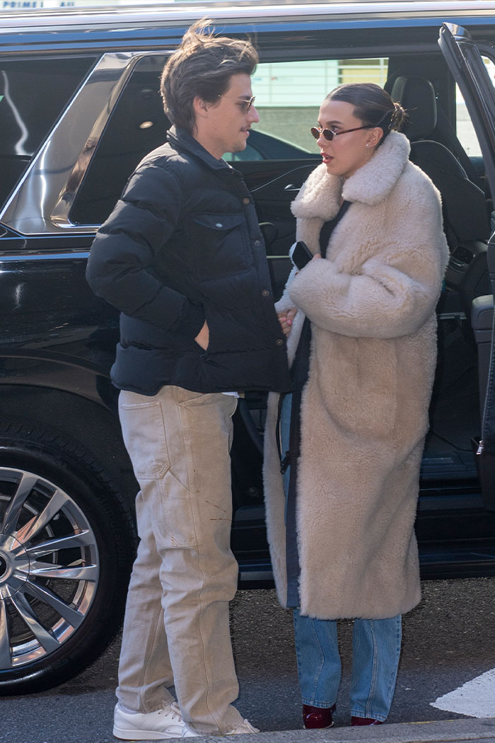 Millie Bobby Brown wearing a long coat and sunglasses, talking to a man beside a black vehicle outdoors.