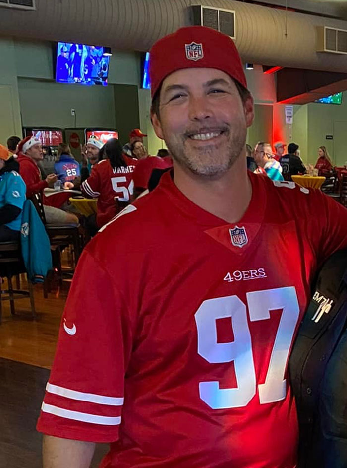 Man in a red NFL 49ers jersey and cap smiling in a crowded sports bar during a game day gathering. Man in a red NFL 49ers jersey and cap smiling in a crowded sports bar during a game day gathering.