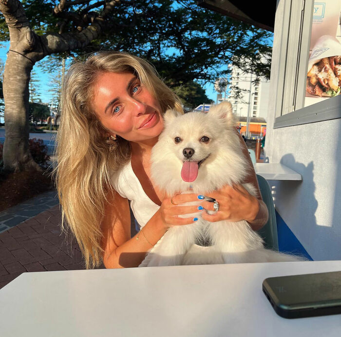 Blonde woman holding a fluffy white dog outdoors in bright sunlight, related to Bonnie Blue controversy in Bali. Blonde woman holding a fluffy white dog outdoors in bright sunlight, related to Bonnie Blue controversy in Bali.