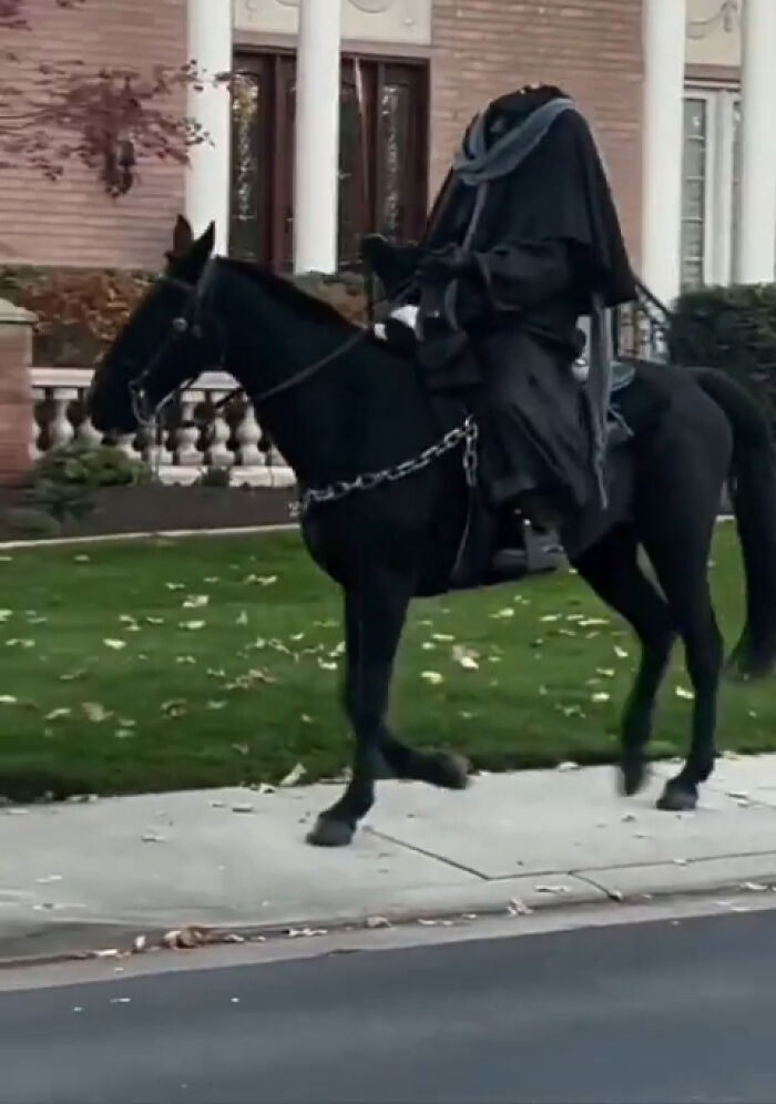 Person dressed in a headless horseman costume riding a black horse on a suburban sidewalk, interesting sight shared online