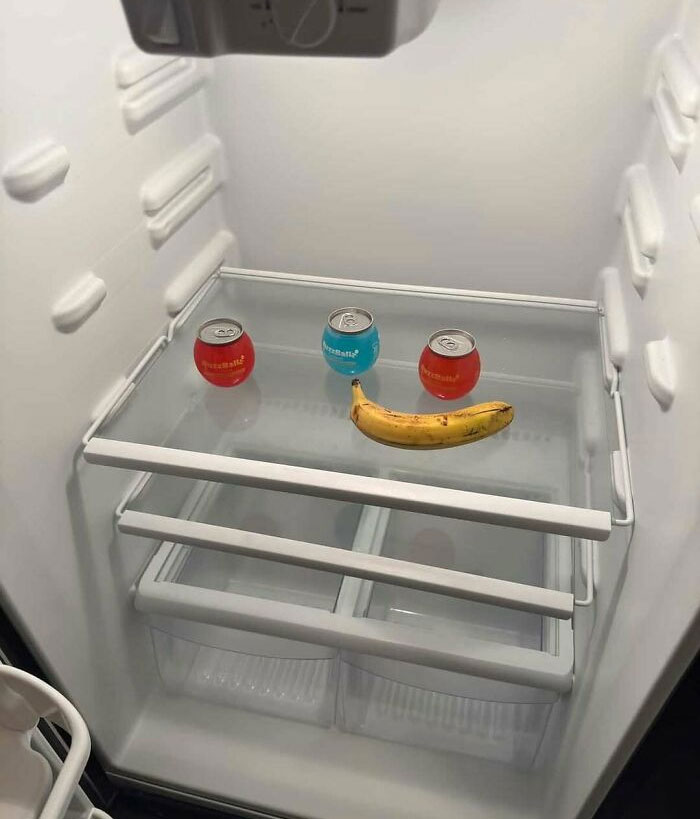 Empty dude fridge with three small drink cans and a banana arranged to resemble a smiling face inside.