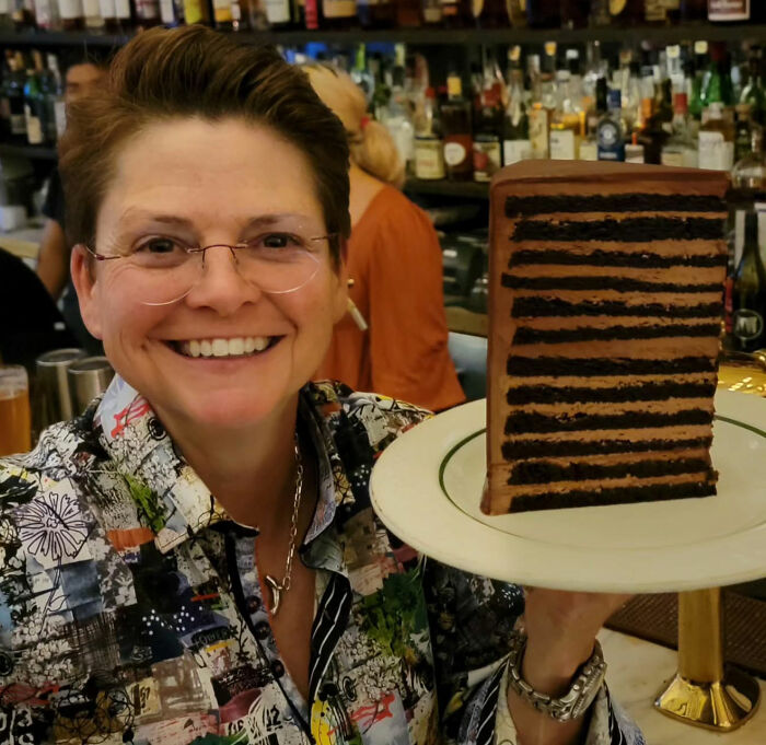 Smiling woman at a bar holding a plate with a large slice of layered chocolate cake, NYC setting. Smiling woman at a bar holding a plate with a large slice of layered chocolate cake, NYC setting.