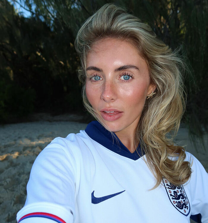 Blonde woman with blue eyes wearing a white England football jersey outdoors, reflecting on Bonnie Blue arrest fate after controversial video.