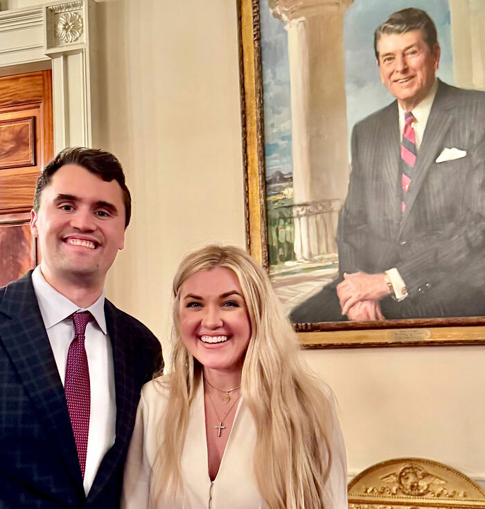 Erika Kirk smiling with a male companion indoors at a TPUSA event, with a portrait of Ronald Reagan in the background. Erika Kirk smiling with a male companion indoors at a TPUSA event, with a portrait of Ronald Reagan in the background.