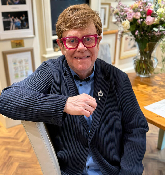 Elton John wearing red glasses and a navy jacket indoors, reacting to fans trolling him over a kitchen detail. Elton John wearing red glasses and a navy jacket indoors, reacting to fans trolling him over a kitchen detail.