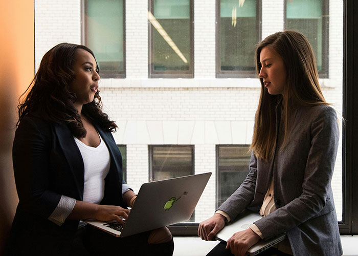 Two coworkers discussing work by the window with laptops, capturing workplace tension and HR involvement themes. Two coworkers discussing work by the window with laptops, capturing workplace tension and HR involvement themes.