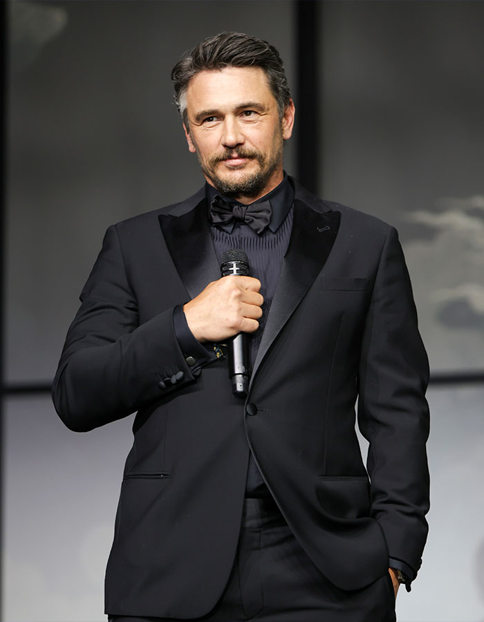 James Franco in a black tuxedo holding a microphone, speaking at an event, reflecting on being cast out of Hollywood.