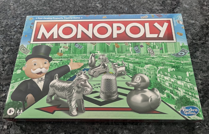 Monopoly board game box with classic playing pieces including dog, thimble, duck, and car on a green city background.