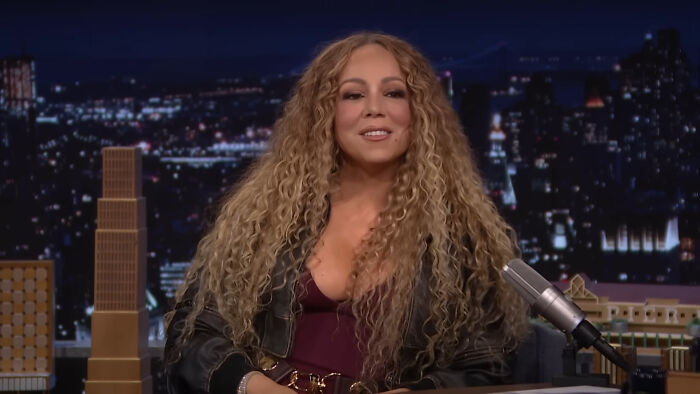 Singer with long curly hair sharing fascinating secrets about celebrities in a late-night talk show interview.