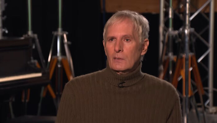 A man in a brown turtleneck sweater sitting in a dimly lit studio, related to celebrities people think suffer.