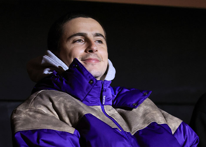 Man wearing a purple and beige jacket, smiling slightly, in a dark indoor setting related to Timothée Chalamet arrogance controversy. Man wearing a purple and beige jacket, smiling slightly, in a dark indoor setting related to Timothée Chalamet arrogance controversy.