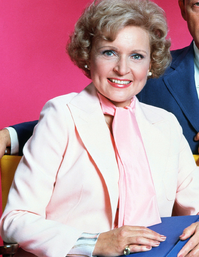 Female surgeon smiling in a light pink suit and scarf seated against a bright pink background in a studio setting.