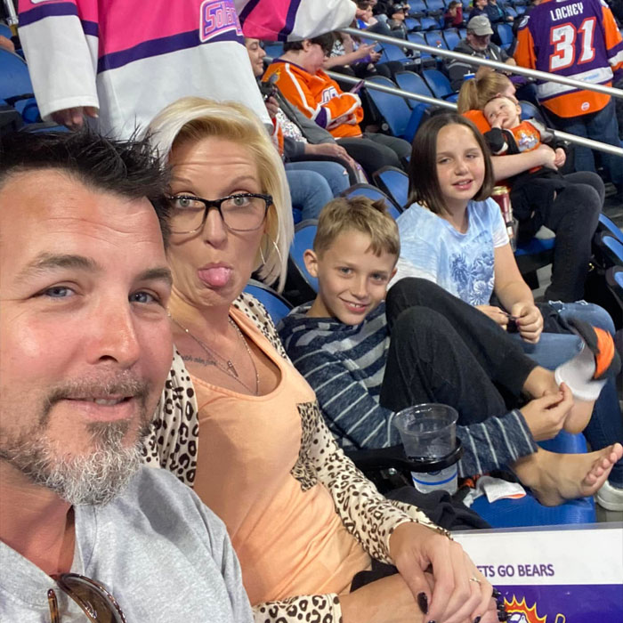Family at NFL game, man in foreground with beard, woman making a face, kids smiling in stadium seats.