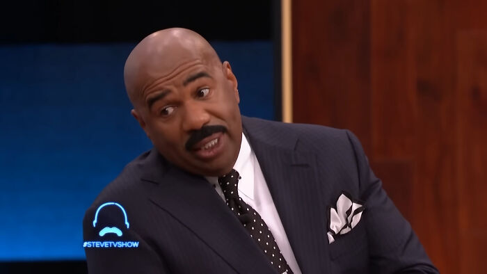 Steve Harvey on a talk show set, wearing a dark suit and polka dot tie, discussing people think celebrities closet skeletons.