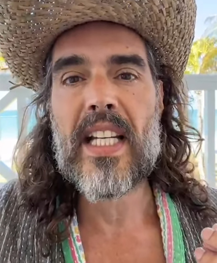 Russell Brand speaking outdoors wearing a straw hat and patterned shirt, showing a bitter reaction to ex-wife Katy Perry news.