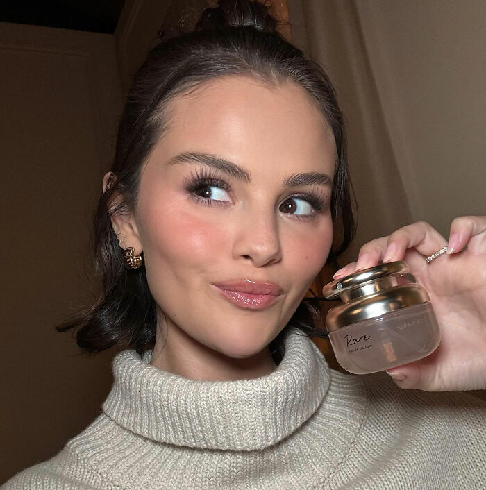 Selena Gomez holding Rare beauty product, smiling and looking to the side with flawless makeup and styled hair indoors. Selena Gomez holding Rare beauty product, smiling and looking to the side with flawless makeup and styled hair indoors.