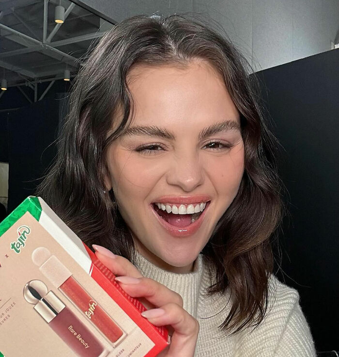 Selena Gomez smiling and holding a Rare Beauty lip gloss product in a casual indoor setting Selena Gomez smiling and holding a Rare Beauty lip gloss product in a casual indoor setting