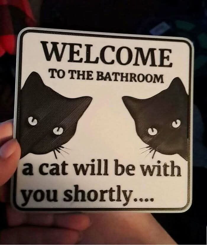 Hand holding a bathroom sign with two black cat illustrations and text about a cat being with you shortly.