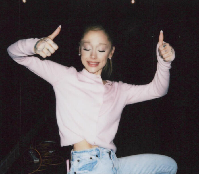 Ariana Grande wearing a light pink sweater and jeans, giving thumbs up with eyes closed against a black background. Ariana Grande wearing a light pink sweater and jeans, giving thumbs up with eyes closed against a black background.