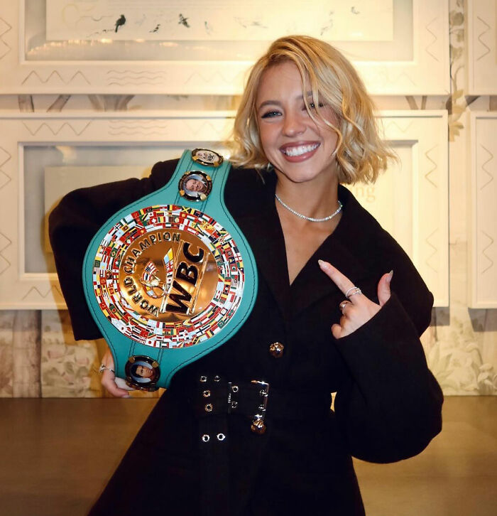 Sydney Sweeney smiling and holding a colorful WBC championship belt indoors in a casual black outfit. Sydney Sweeney smiling and holding a colorful WBC championship belt indoors in a casual black outfit.