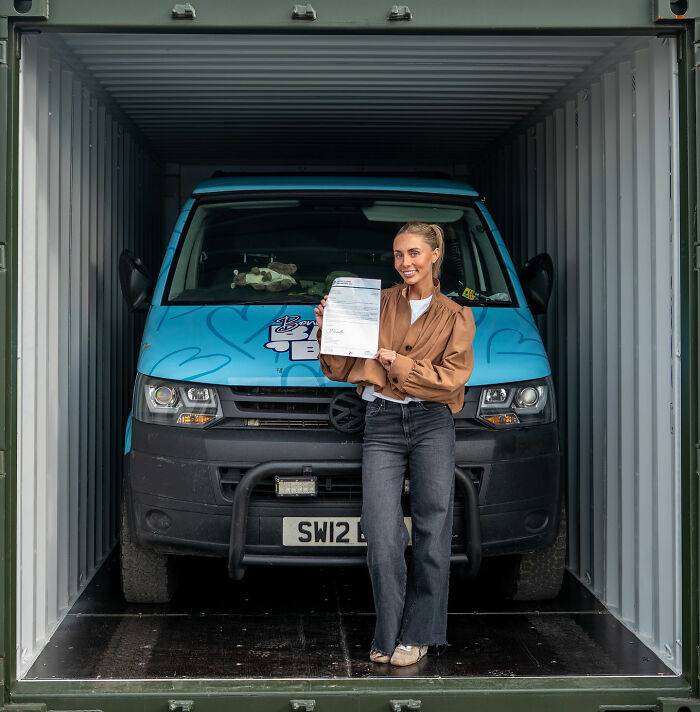 Woman standing inside a container holding a document with a blue Bonnie Blue van behind her, related to Bonnie Blue controversy in Bali. Woman standing inside a container holding a document with a blue Bonnie Blue van behind her, related to Bonnie Blue controversy in Bali.