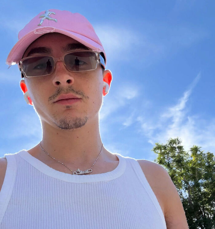 Young man wearing a pink cap and sunglasses, posing outdoors under a clear blue sky, showing a casual red carpet look. Young man wearing a pink cap and sunglasses, posing outdoors under a clear blue sky, showing a casual red carpet look.