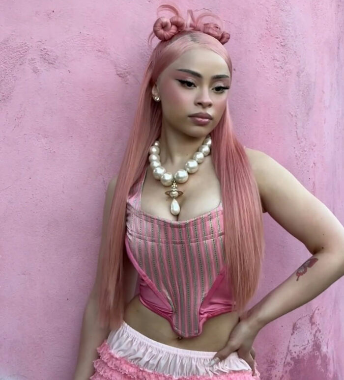 Ice Spice posing against a pink wall wearing a pink corset and pearl necklace sparking outfit outrage among fans. Ice Spice posing against a pink wall wearing a pink corset and pearl necklace sparking outfit outrage among fans.