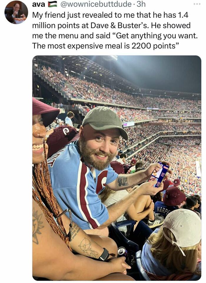 Man smiling at a crowded stadium showing his phone, featured in cheerful memes about wasting time online.