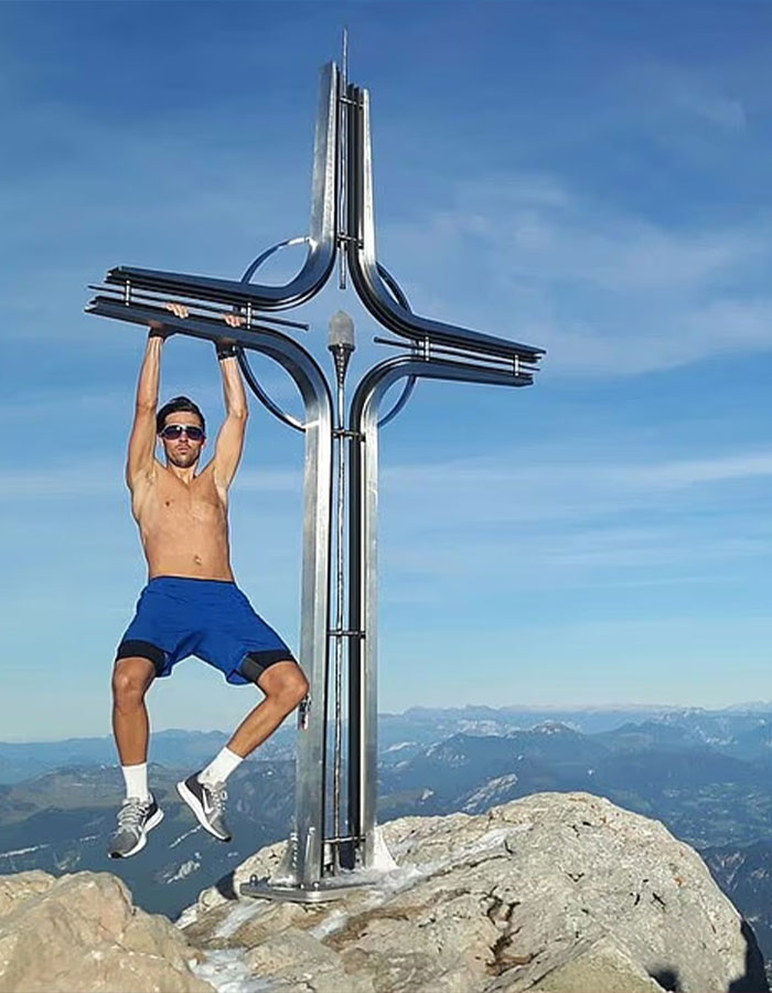 Man wearing sunglasses and blue shorts hanging from metal cross on Austria’s highest peak with mountain landscape behind him Man wearing sunglasses and blue shorts hanging from metal cross on Austria’s highest peak with mountain landscape behind him