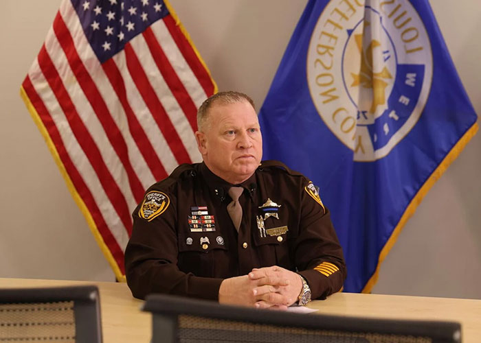Law enforcement officer sitting at a table with American and county flags, discussing child kidnapped from Kentucky found alive case. Law enforcement officer sitting at a table with American and county flags, discussing child kidnapped from Kentucky found alive case.