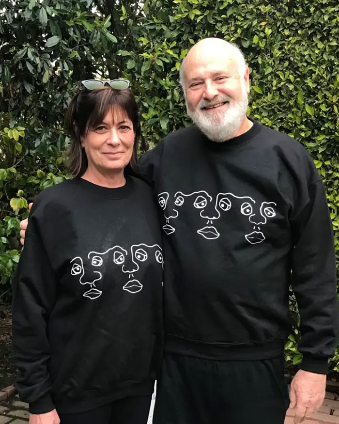 Rob Reiner smiling outdoors with a woman, both wearing matching black sweatshirts with abstract face designs.