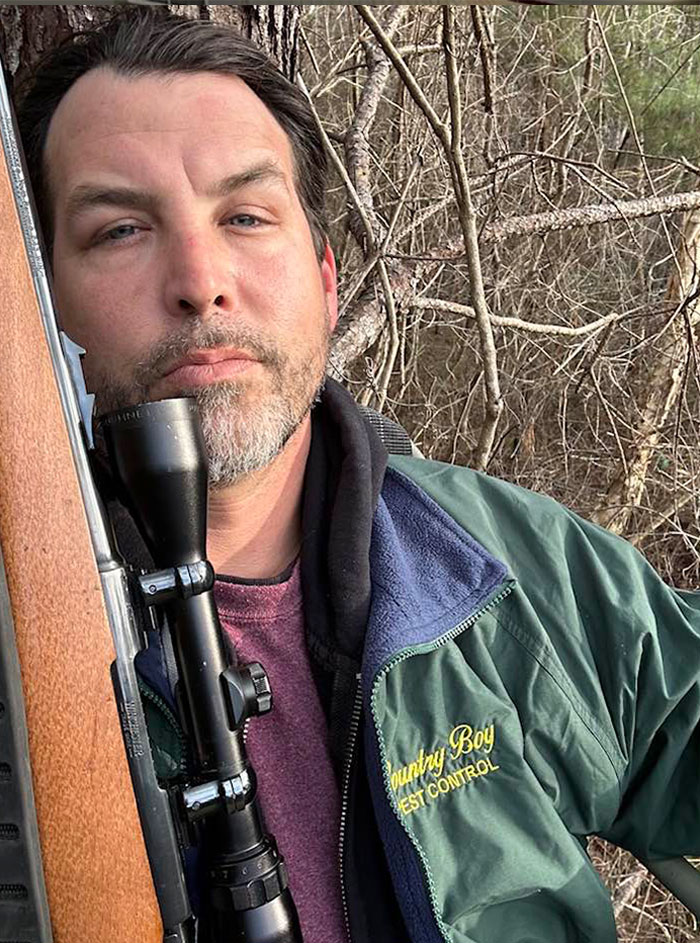 Man outdoors holding rifle with scope, wearing green jacket, amid bare tree branches in a tense NFL argument context. Man outdoors holding rifle with scope, wearing green jacket, amid bare tree branches in a tense NFL argument context.