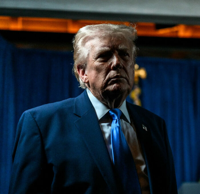 Donald Trump in a dark suit and blue tie, standing in front of blue curtains during a dimly lit event. Donald Trump in a dark suit and blue tie, standing in front of blue curtains during a dimly lit event.