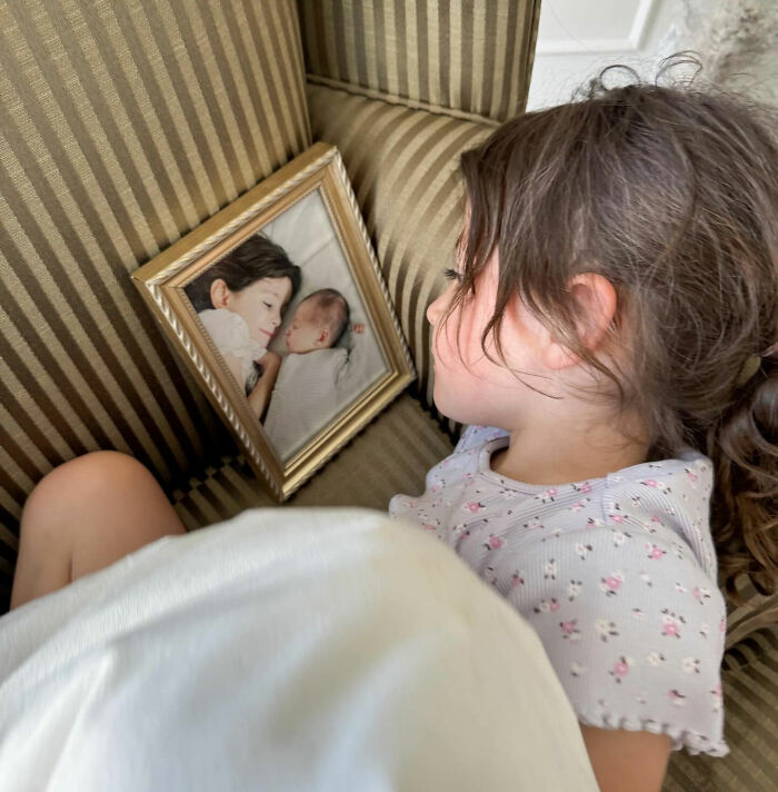Young girl lying on a striped couch holding a framed photo of two sleeping children, reflecting on loss of influencer's daughter. Young girl lying on a striped couch holding a framed photo of two sleeping children, reflecting on loss of influencer's daughter.