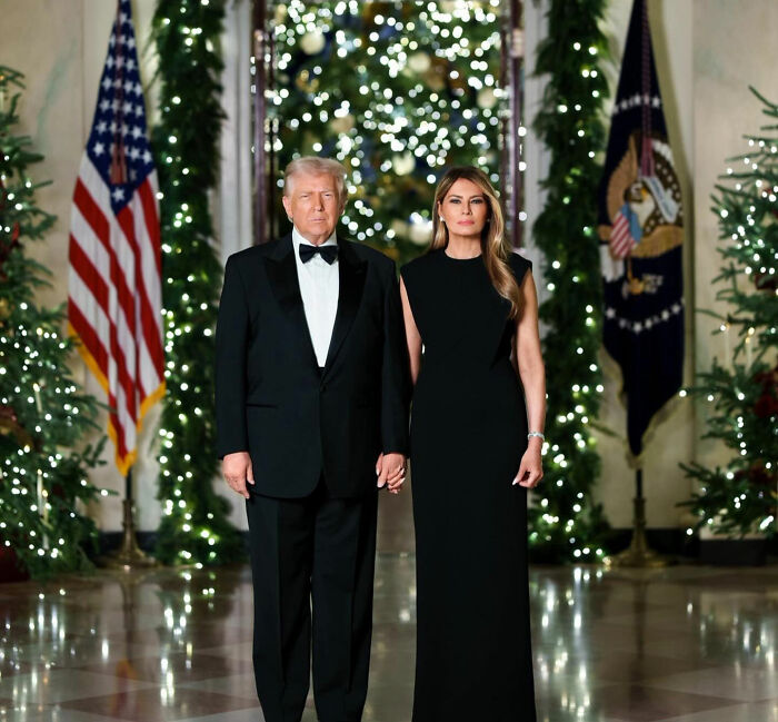 Donald Trump and Melania Trump in formal attire standing by Christmas trees with festive lights during holiday event. Donald Trump and Melania Trump in formal attire standing by Christmas trees with festive lights during holiday event.