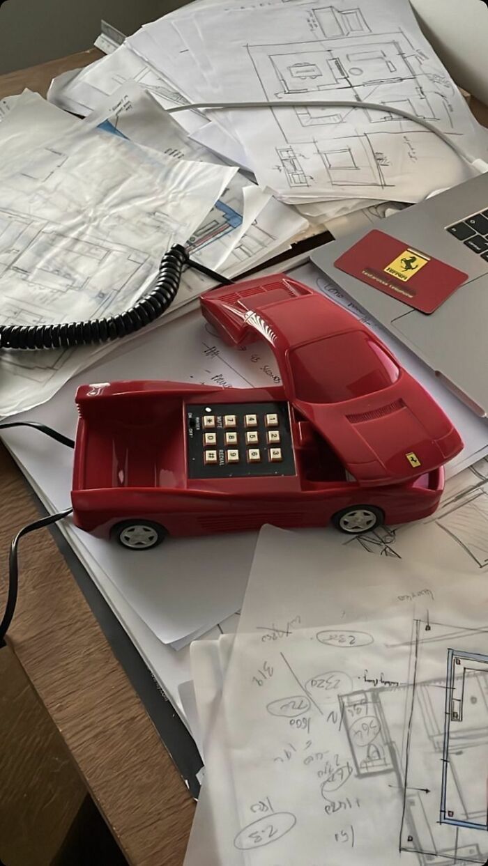 Red car-shaped phone with keypad on a cluttered desk of architectural sketches in a trippy pictures setting.